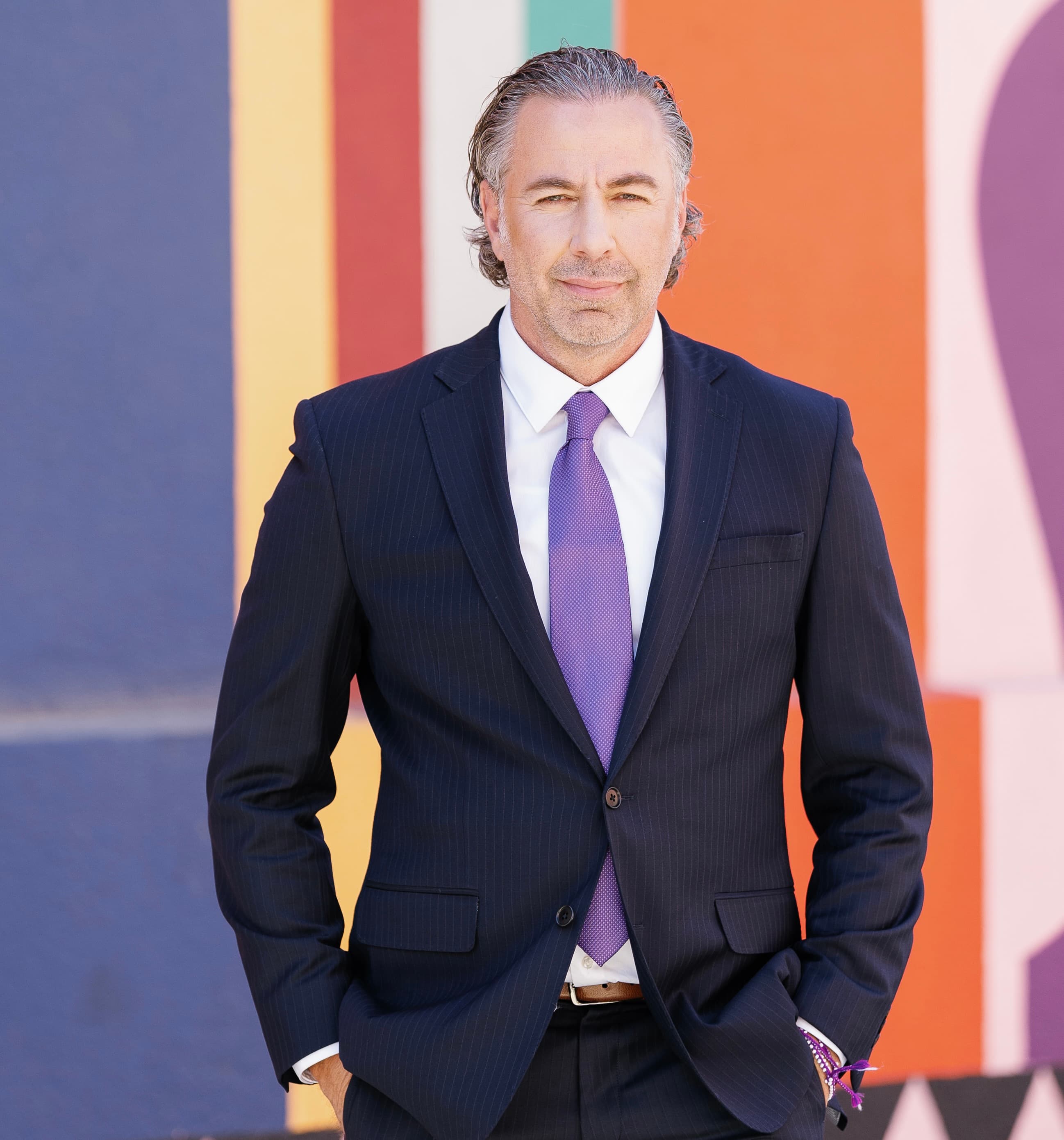 Middle-aged man in a pinstripe suit and purple tie against a colorful striped background.